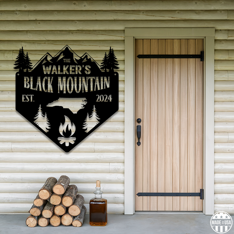 A customizable black metal sign mounted on a wooden wall, featuring a door, logs, and a jug with moonshine with text that reads 'Black Mountain Est. 2024.'