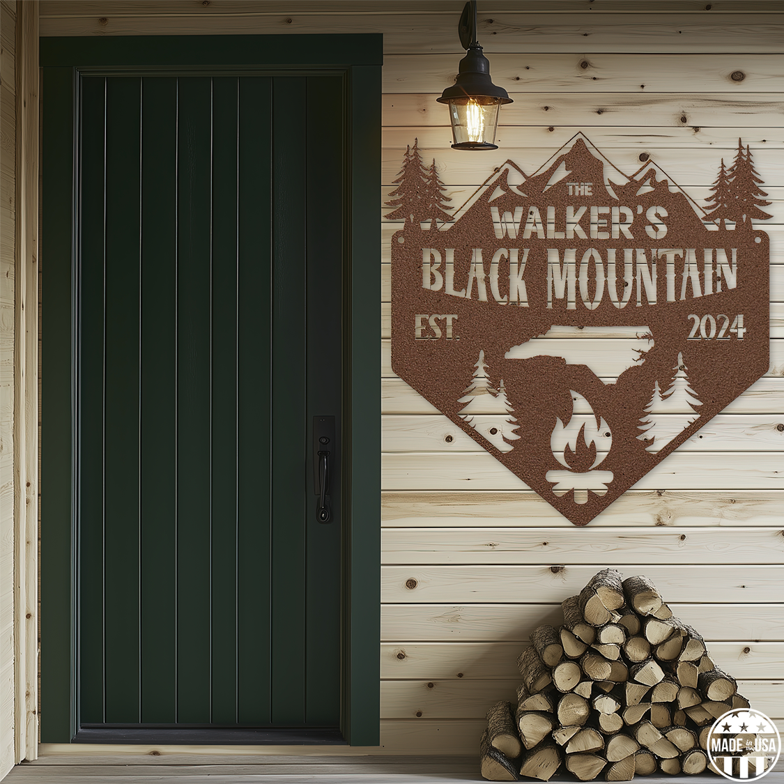 A customizable copper metal sign mounted on a wooden wall, featuring a mountain, trees, and a campfire with text that reads 'Black Mountain Est. 2024.' The sign is beside a green door, and chopped logs are beneath it.