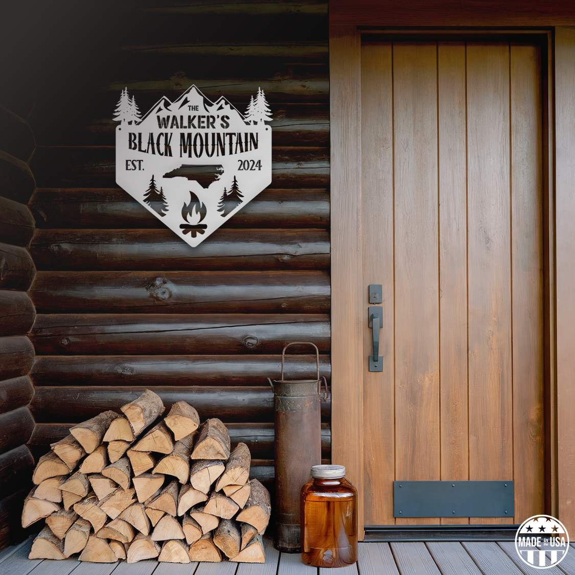 A customizable white metal sign mounted on a cabin wall, featuring a mountain, trees, and a campfire with text that reads 'Black Mountain Est. 2024.' There is chopped wood outside the cabin door.