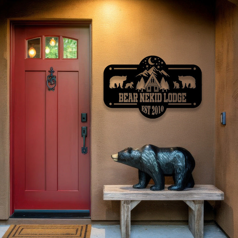 A cozy lodge entrance with a red wooden door features a custom black metal sign reading 'BEAR NEKID LODGE EST 2010,' mounted on the wall. The sign showcases silhouettes of bears and a cabin under a starry night sky with pine trees. A wooden bench sits below the sign, adorned with a carved black bear statue. The warm-toned stucco walls and rustic decor create a welcoming cabin atmosphere.