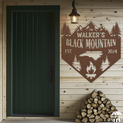 A customizable copper metal sign mounted on a wooden wall, featuring a mountain, trees, and a campfire with text that reads 'Black Mountain Est. 2024.' The sign is beside a green door, and chopped logs are beneath it. 