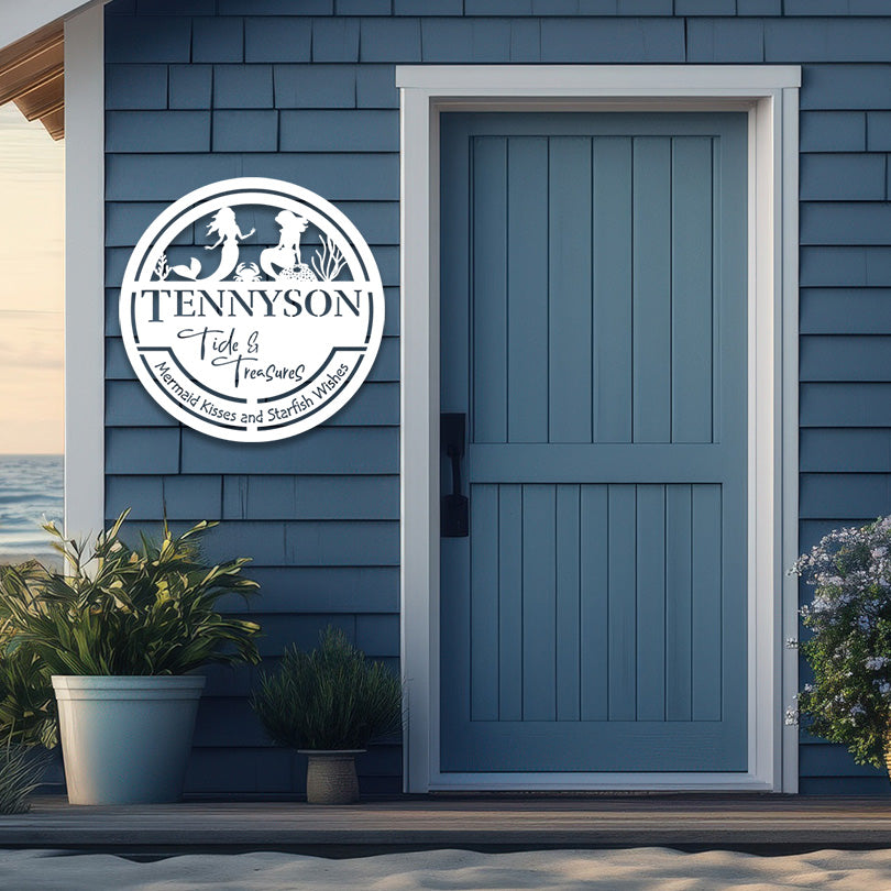 A beach house with blue siding and a matching blue door features a personalized round metal sign mounted beside the entrance. The sign reads 'TENNYSON Tide & Treasures' with the tagline 'Mermaid Kisses and Starfish Wishes,' and includes silhouettes of a mermaid and beach elements. Two potted plants are placed near the door, and the beach shoreline is visible in the background, enhancing the coastal ambiance.