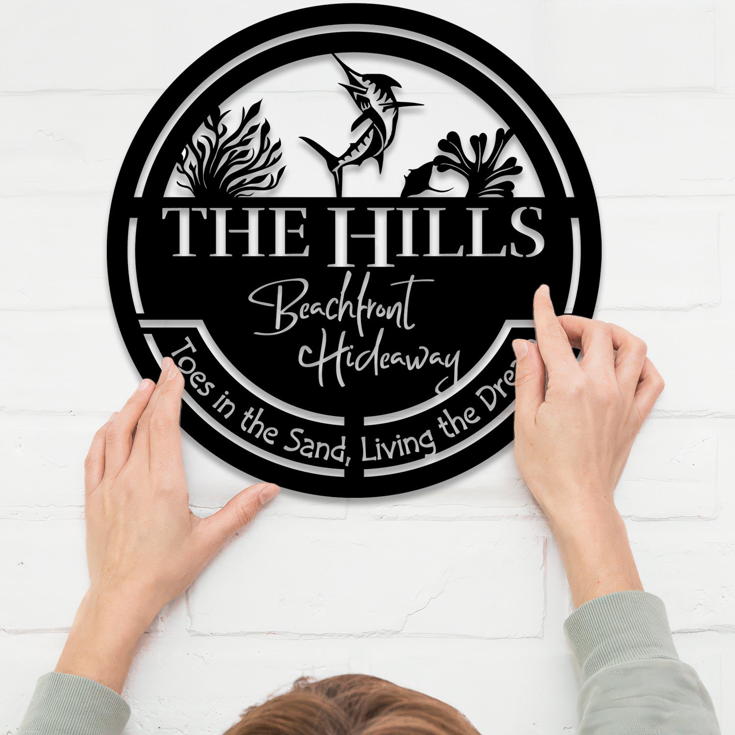 A person holding a black metal sign that reads 'THE HILLS Beachfront Hideaway' with the phrase 'Toes in the Sand, Living the Dream.' The sign is mounted on a white brick wall, showcasing its durability and elegant design. The accompanying text highlights the sign's craftsmanship, made from 18-gauge steel with a powder-coated finish, ensuring long-lasting quality for both indoor and outdoor use. This image emphasizes the product's resilience and timeless appeal, perfect for enhancing any space.