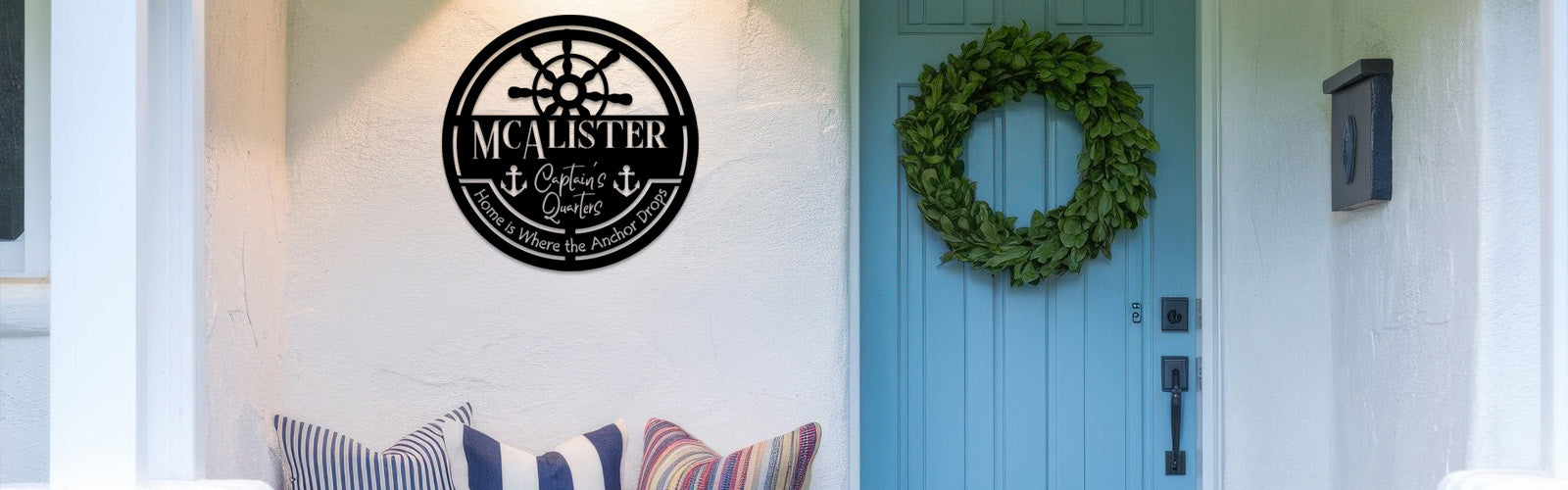 A charming coastal-themed front porch featuring a personalized black metal sign that reads 'MCALISTER Captain's Quarters' with the phrase 'Home is Where the Anchor Drops' and nautical elements such as a ship's wheel and anchors. The sign is mounted beside a bright blue front door adorned with a lush green wreath. The cozy seating area includes striped and solid-colored pillows, creating a welcoming and nautical ambiance. The white stucco walls and decorative elements enhance the coastal charm of the home.